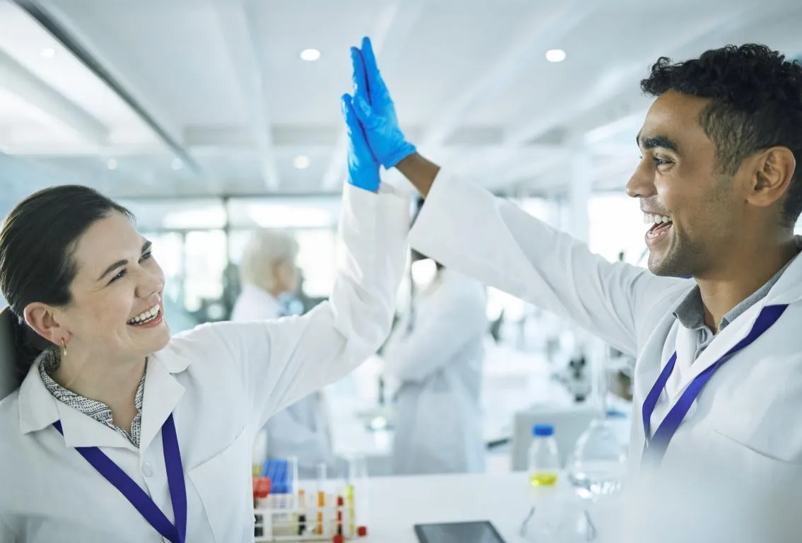 Dos científicos con batas de laboratorio, uno con guantes azules, chocando las manos en un entorno de laboratorio, simbolizando una colaboración exitosa o un avance.