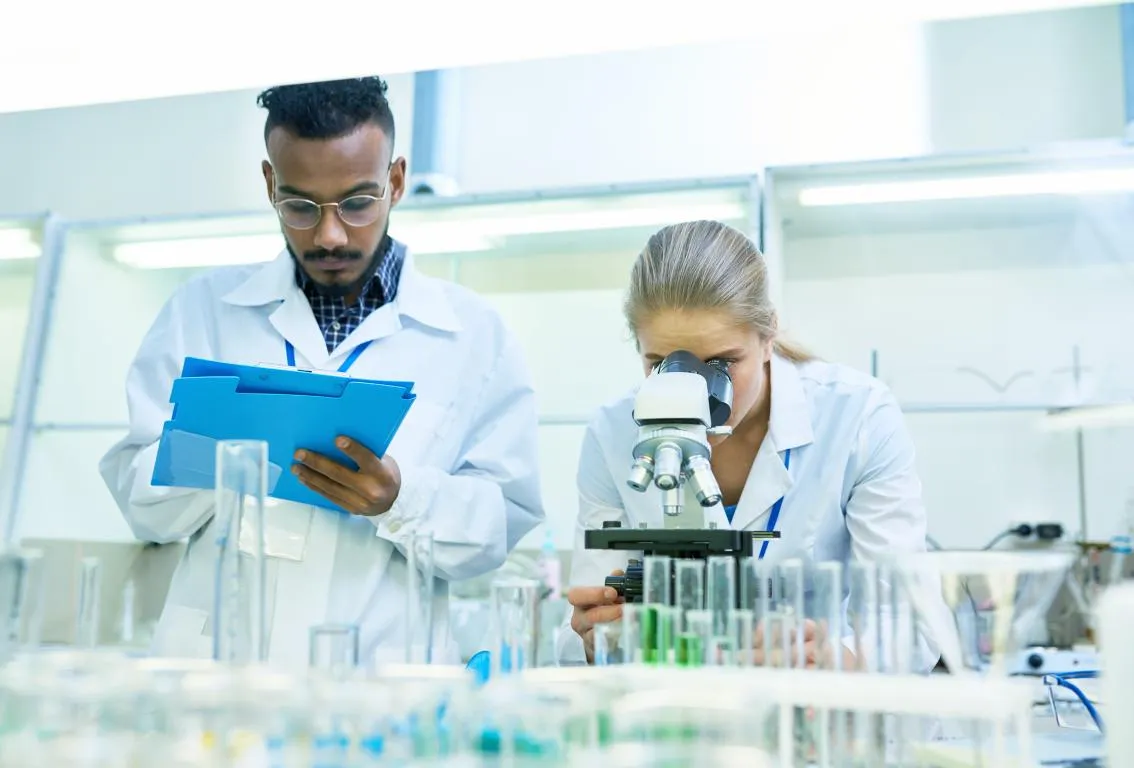 Dos científicos, un hombre y una mujer, con batas de laboratorio y gafas de seguridad, trabajando con equipos de laboratorio, lo que representa investigación científica y colaboración.