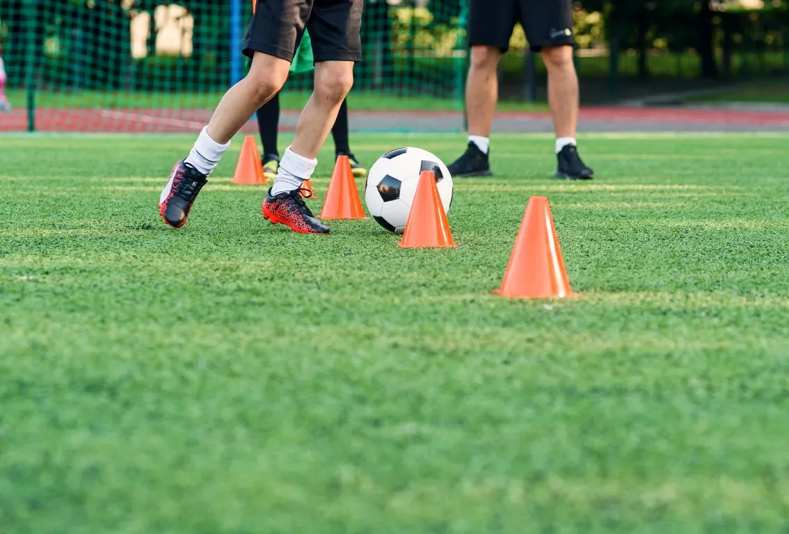Una toma de cerca de las piernas de futbolistas en un campo verde con un balón y conos de entrenamiento naranjas, representando metodología ágil, entrenamiento o preparación estratégica.