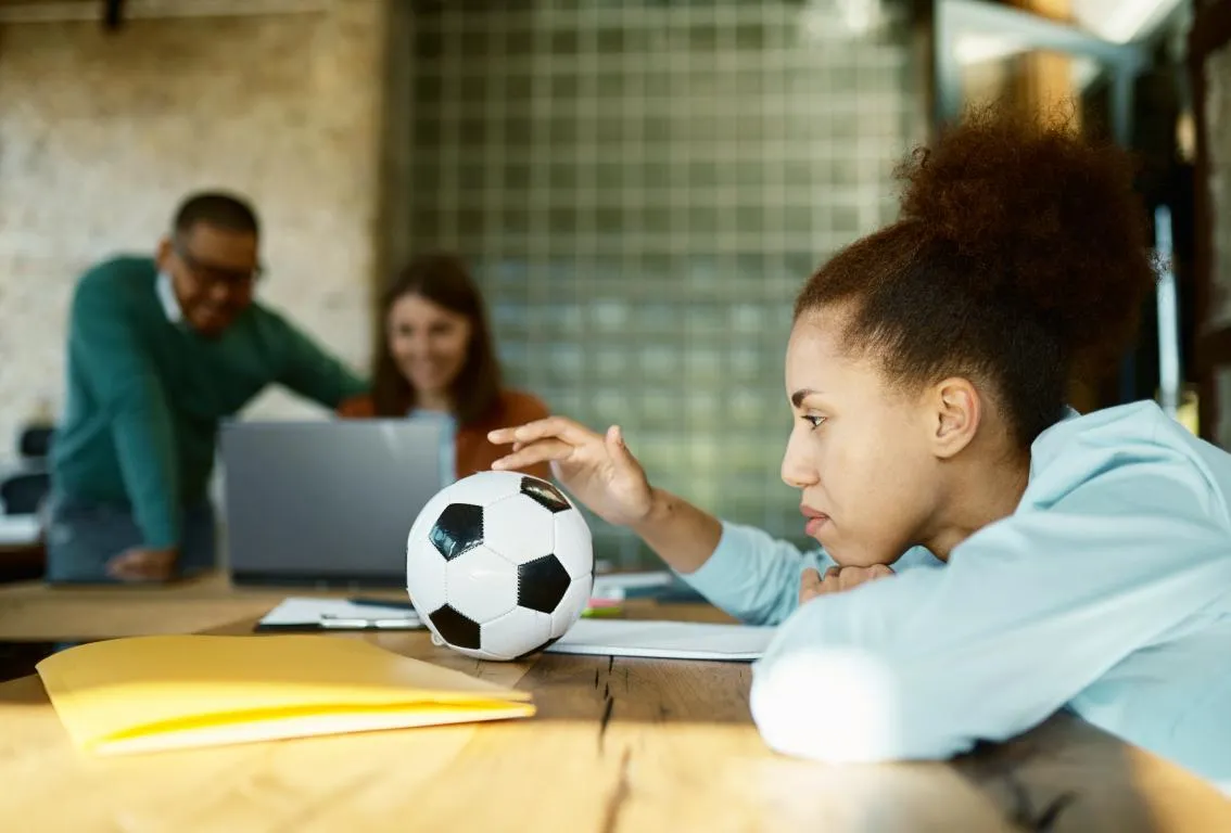 Un grupo diverso de personas colaborando alrededor de una mesa en una oficina, con un balón de fútbol sobre la mesa, simbolizando trabajo en equipo, estrategia dinámica o innovación.