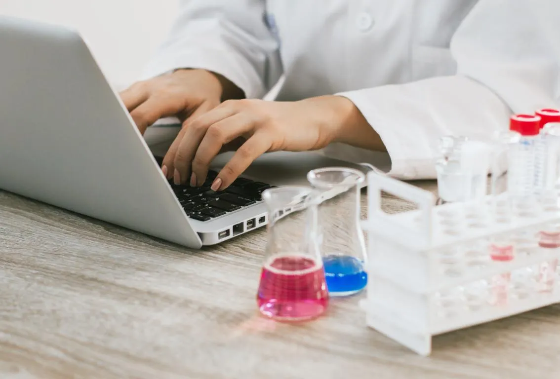 Un primer plano de manos tecleando en un teclado de portátil, con material de laboratorio que contiene líquidos de colores en el fondo, lo que sugiere investigación científica, análisis de datos o tecnología en un entorno de laboratorio.
