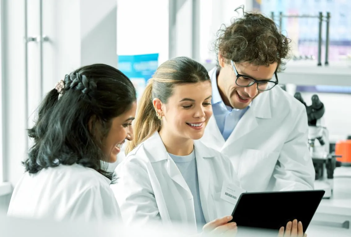 Tres científicos con batas de laboratorio colaborando y observando una muestra o datos, lo que representa trabajo en equipo e investigación en un entorno de laboratorio.