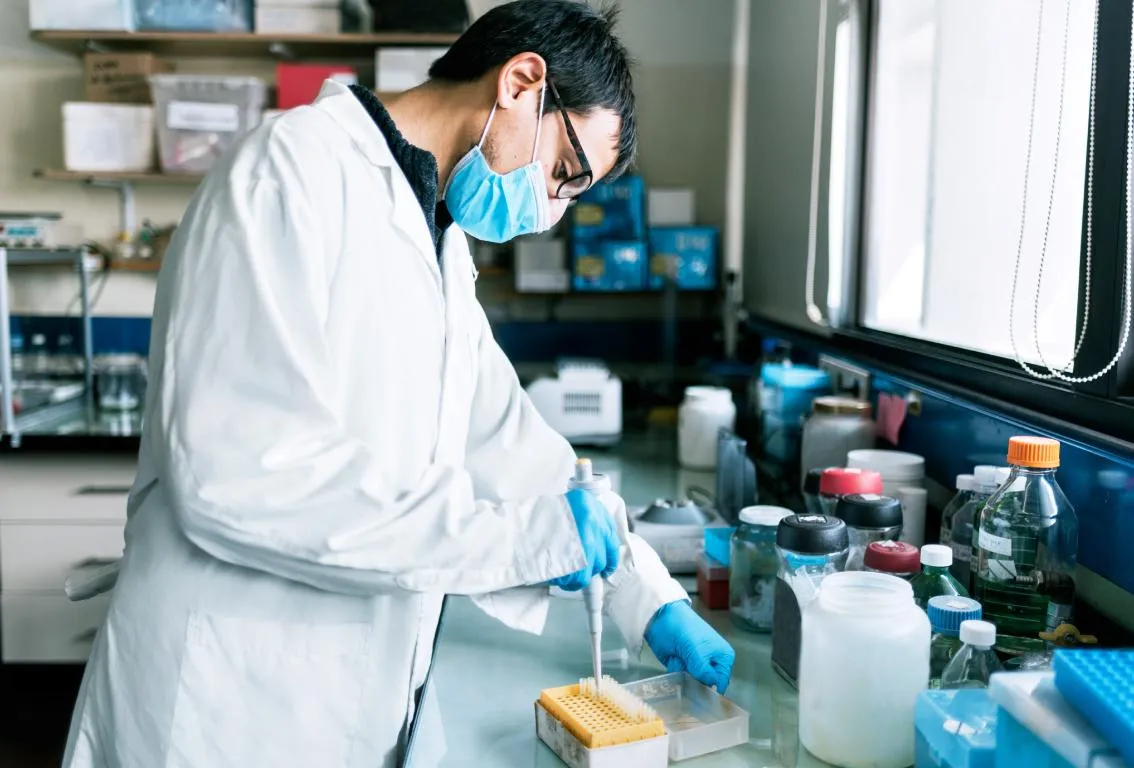 Un científico con bata de laboratorio, guantes y mascarilla, trabajando meticulosamente con vasos de precipitados y muestras en un entorno de laboratorio, simbolizando análisis científico o control de calidad.